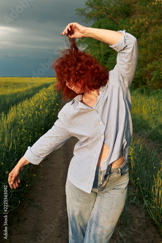 A red-haired woman poses playfully on a rural path beside green crops.
