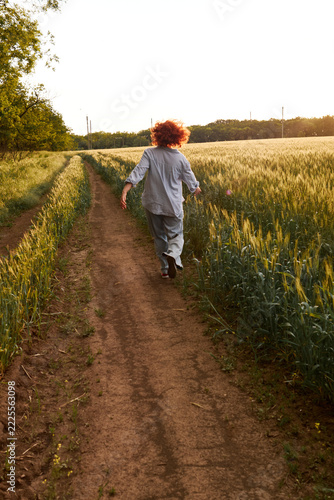 Red-haired woman walks along a dirt path between sunlit golden grain fields