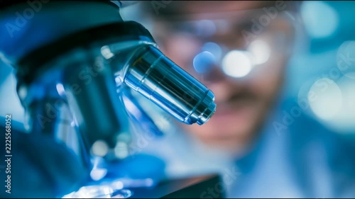 Microscope objective lenses fill the foreground as a technician wearing protective goggles works behind them in a laboratory with cool blue lighting