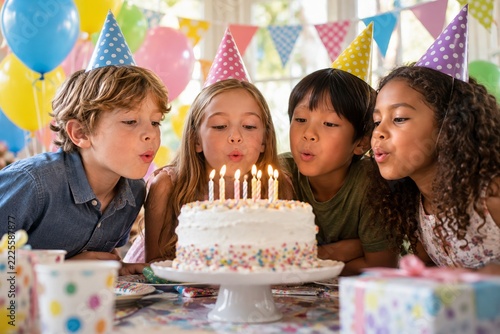 Children in colorful party hats blowing birthday candles on white cake