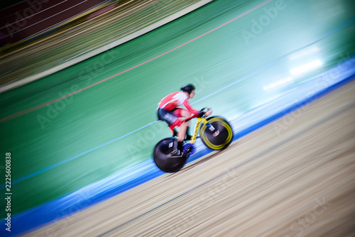 The photograph captures a cyclist speeding along the steeply banked track of a velodrome. The athlete is wearing a red-and-white racing suit and a helmet. Leaning low over the handlebars of an aerodyn