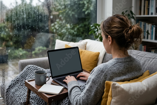 Woman working remotely on a laptop on the sofa on a rainy day, over-the-shoulder view