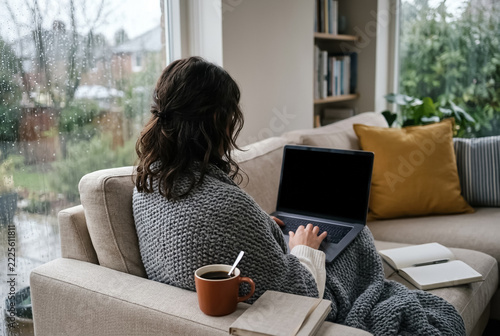 Woman working remotely on a laptop on the sofa on a rainy day, over-the-shoulder view