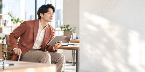 窓から陽光が差し込む開放的なオフィスで タブレットを手に立ち上がろうと微笑むビジネスマン テラコッタ色のジャケットと右側の広いコピースペース