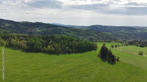 Elevated perspective shows winding roads through green fields, wooded hills, and open meadows, ending at small village on calm horizon beneath overcast clouds.