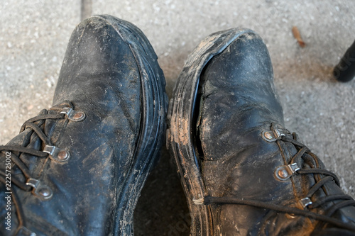 Pair of heavily worn, weathered, and caked-in-mud black leather military boots. Old, cracked and battered work boots covered in dried mud and deep creases from heavy labor. Rugged utility boots
