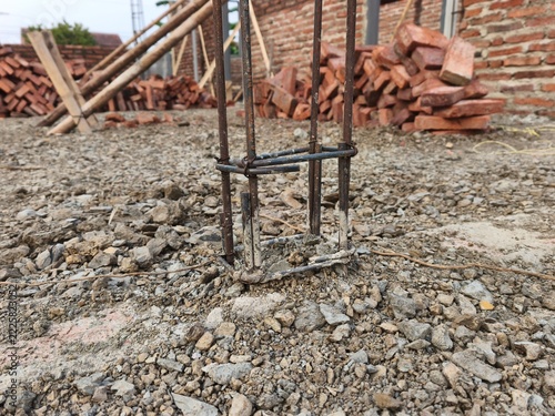 Steel rebar column reinforcement emerging from gravel ground on active house construction site with red bricks pile in background, ideal for civil engineering and structural building photo.