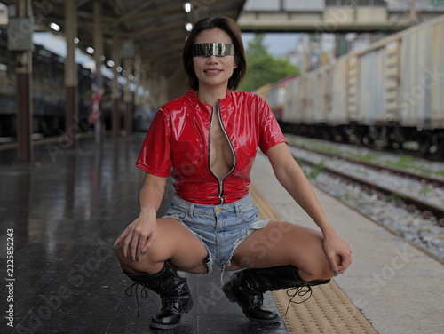 A woman wearing futuristic metallic eyewear, an unzipped red vinyl top, denim shorts, and black boots crouches on a train station platform