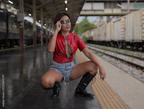 A woman wearing metallic eyewear, a red vinyl top, denim shorts, and black boots crouches on a train station platform