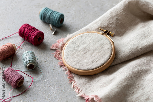 Soft linen in wooden hoop rests beside colorful thread spools. Frustrated yarn strands lie tangled on neutral concrete surface. Warm tones suggest calm, creative crafting moment. Ideal for home decor