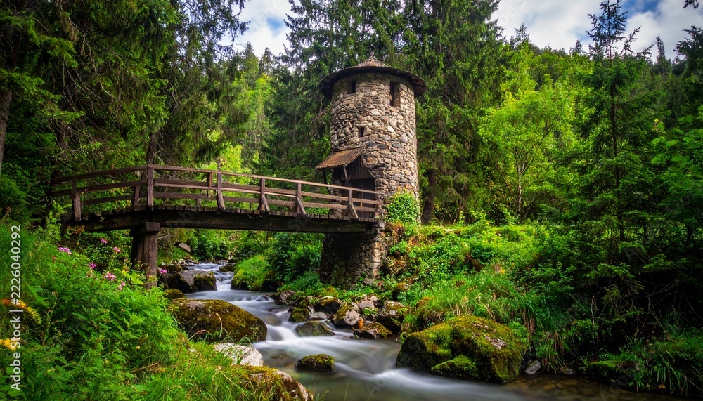 Fototapeta premium An enchanting stone tower standing beside a flowing river in a lush green forest landscape with a wooden bridge crossing the mountain stream.