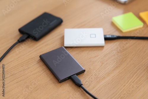 Three external portable hard drives are connected with cables on a wooden office desk. A person uses these data storage devices for digital backups and file transfer work.
