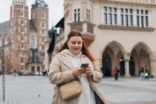 Attractive young female tourist is exploring new city. Redhead girl holding a smart phone on Market Square in Krakow. Traveling Europe in summer. St. Marys Basilica. The Cloth Hall, Vacation concept