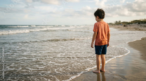 Young boy standing on a sandy beach looking out at the ocean waves under a cloudy sky