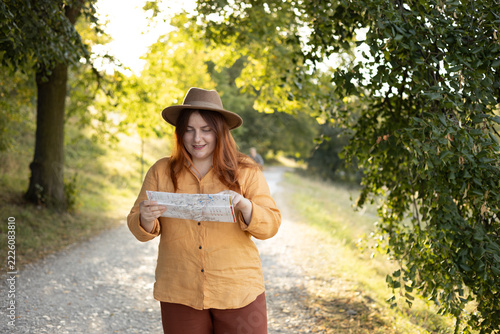 Stylish 30s woman holding paper map relaxing in nature. Travel and active lifestyle concept. Happy female lifestyle wearing backpack and hat exercising outdoors.