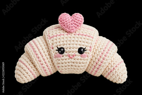 Handmade amigurumi crochet character of a cute kawaii croissant with a happy face and pink heart on top, isolated on black background for Valentine's Day or bakery cafe branding.