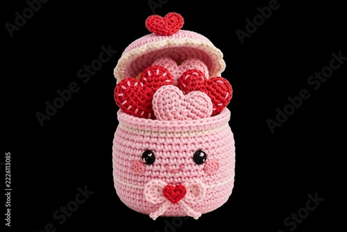 Amigurumi crochet craft of a charming pink kawaii character basket overflowing with red and pink hearts, isolated on a black background for a sweet Valentine's Day gift concept.