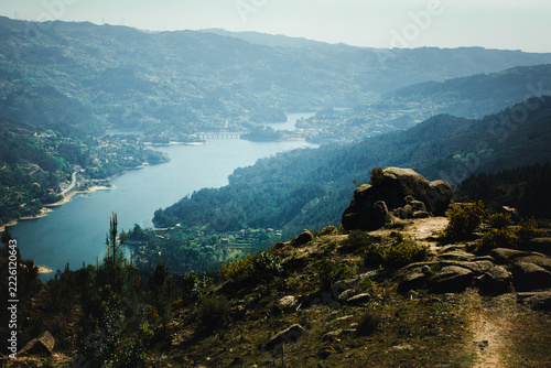 Panoramic view of a mountain reservoir winding through a forested valley with rocky foreground and a distant bridge