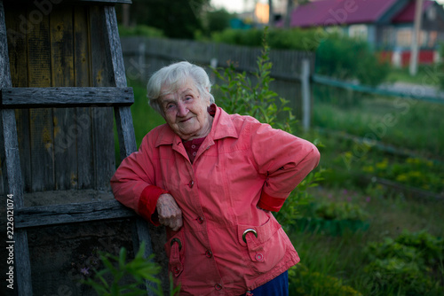 Smiling elderly woman leaning on a weathered wooden ladder beside a rustic house in a garden