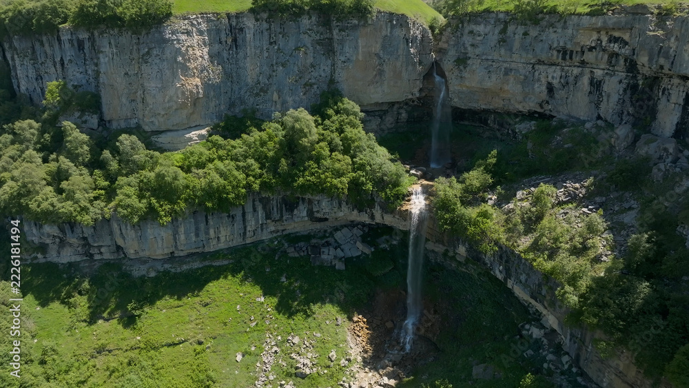 Fototapeta premium The Matlas waterfalls descend over a pale limestone ledge into a wooded ravine, where clear water winds among boulders and summer greenery beneath wide skies near Matlas, Dagestan, Russia
