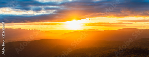 Dramatic storm clouds gather over the rugged mountain ranges of Grampians National Park in Victoria, Australia, as the setting sun casts warm light across the expansive landscape.