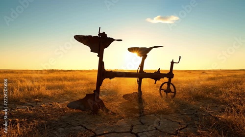Silhouette of an old rusty vintage plow standing in a dry cracked field at sunset with golden light