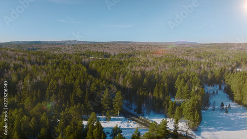 An expansive aerial view reveals a snow-covered conifer wilderness and winding frozen waterway beneath soft winter light, showcasing the quiet, remote terrain of the Ural Mountains in Russia