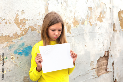 Girl in yellow shirt holding blank paper against a worn wall