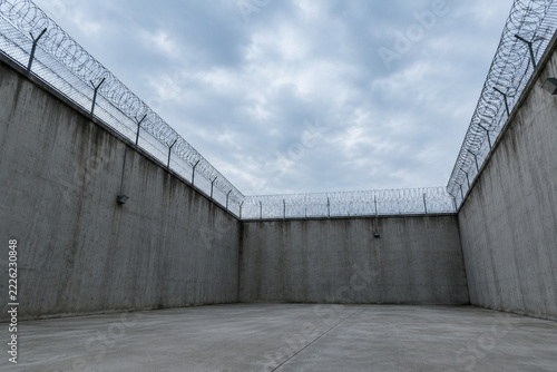 Empty high-walled prison yard with concrete ground and razor wire fences, bleak view.