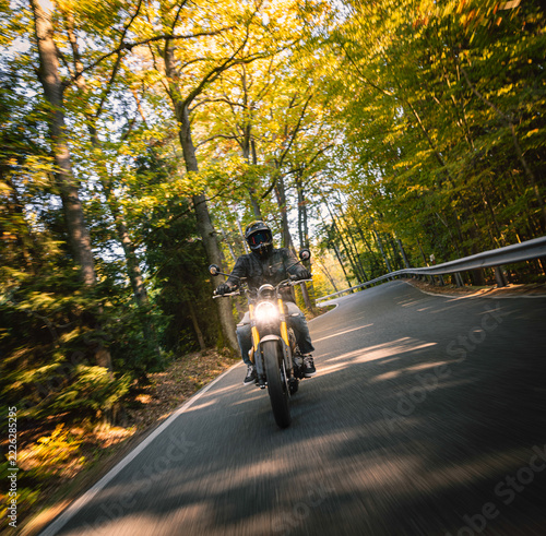 Motorcyclist wearing a black helmet and dark riding gear approaching the camera on a winding asphalt road through a sunlit green and golden autumn forest, with dynamic motion blur.