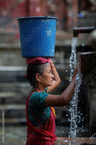 Water flows freely as a woman balances a blue bucket on her head while engaged in a daily ritual at the local fountain during the sunny afternoon