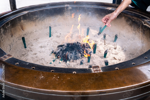 A hand lighting an incense stick from a brazier filled with fire and ash in a Japanese temple on an August day.