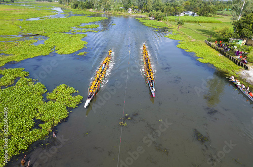 Aerial view of two long traditional boats racing on a river filled with water hyacinths, with rowers in yellow shirts paddling in unison in Rangpur, Rangpur Division, Bangladesh.