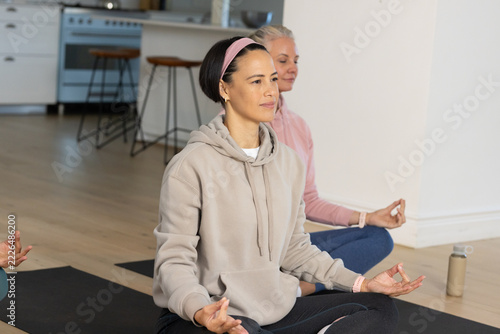 Female friends sitting cross-legged meditating in open-plan living area on black yoga mats
