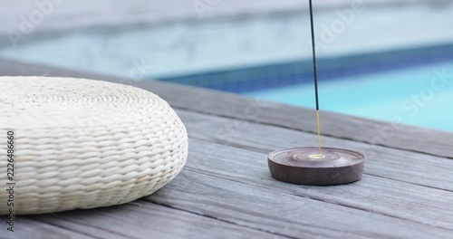 Light breeze is causing incense stick to smolder and curl smoke beside woven cushion for relaxation
