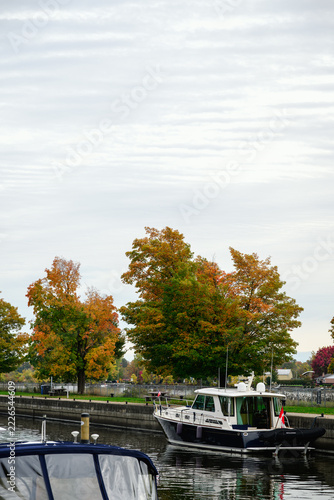 A white motor yacht lies moored at Sainte-Anne-de-Bellevue Canal beneath autumn foliage with a stern flag and reflected hull framed vertically in bright overcast daylight.