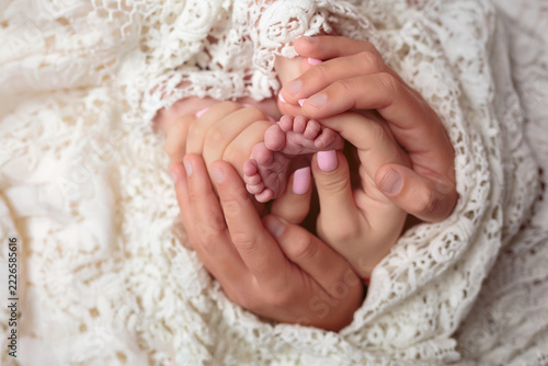 A father holds the mother’s hand, and the mother’s hand holds the infant’s feet, against a white lace background.