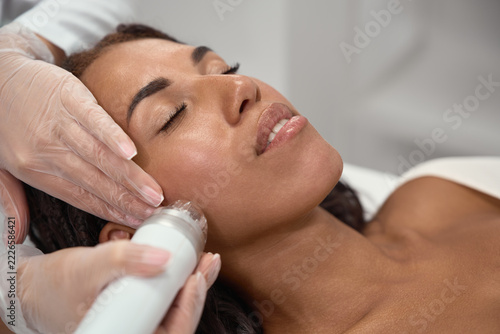 A smiling African woman receives radiofrequency microneedling on her cheek. A cosmetologist applies a needle cartridge during a facial procedure for firmer looking skin.