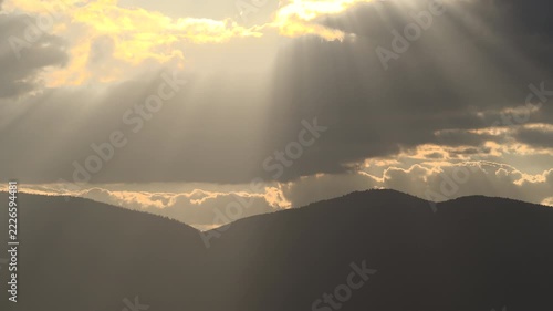 Cinematic time-lapse of dramatic sunbeams breaking through dark clouds over mountain peaks at sunset. Beautiful god rays and golden light illuminating the mountain silhouettes.