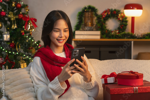 Fotografie Cheerful Asian young woman wearing a white sweater and red scarf, sitting on a sofa and using her smartphone for online messaging during Christmas, festive holiday