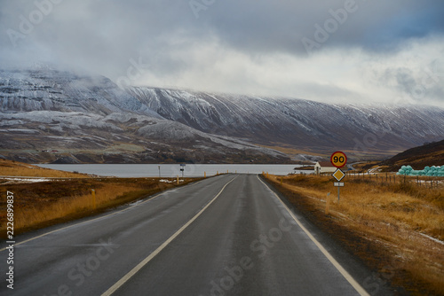 Scenic asphalt highway leading toward fjord and snow-dusted mountains in Iceland. Ring road trip with 90 km speed limit sign during late autumn winter transition.