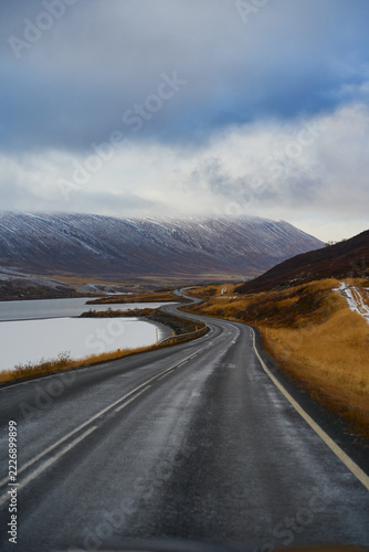 Scenic vertical view of winding asphalt coastal highway along lake toward snow-capped mountain in Iceland. Arctic road trip and self-drive adventure in winter.