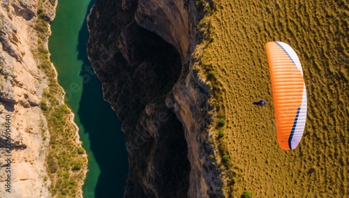Orange paraglider flying over steep canyon with deep green river running through gorge.