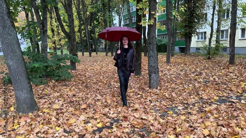 Brunette woman walking with a red umbrella through autumn leaves in a park near residential buildings, rainy weather, seasonal stroll, outdoor lifestyle