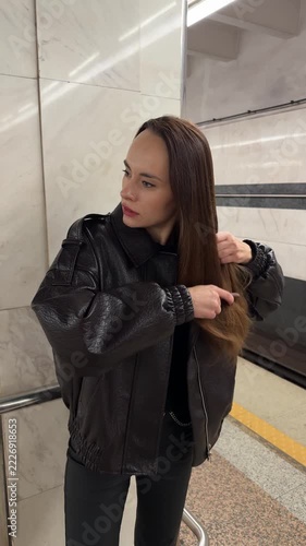Brunette woman combing long brown hair in a tiled subway station wearing a black leather jacket, hair care, daily grooming, urban lifestyle