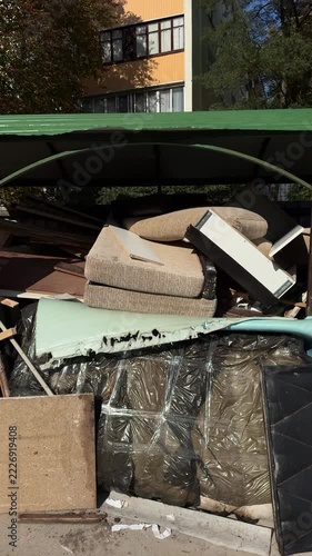 Discarded sofa cushions and broken furniture panels piled under a green canopy beside an apartment building, bulky waste, furniture disposal