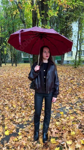 Brunette woman holding a burgundy umbrella among fallen leaves near apartment buildings, autumn scenery, outdoor portrait, casual fashion