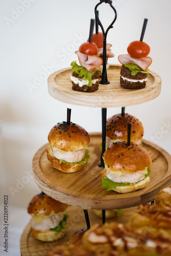 Mini burgers and sandwiches with ham, cheese, vegetables, and greens arranged on a wooden tiered stand for a buffet.