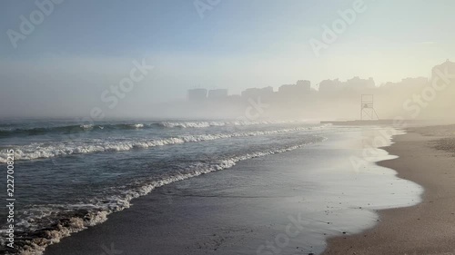 Misty coastal scene with foamy waves, wet sand and silhouetted high rise buildings on hazy horizon