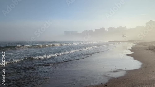 Calm sea waves roll onto sandy beach while city skyline and lifeguard tower fade into thick morning fog